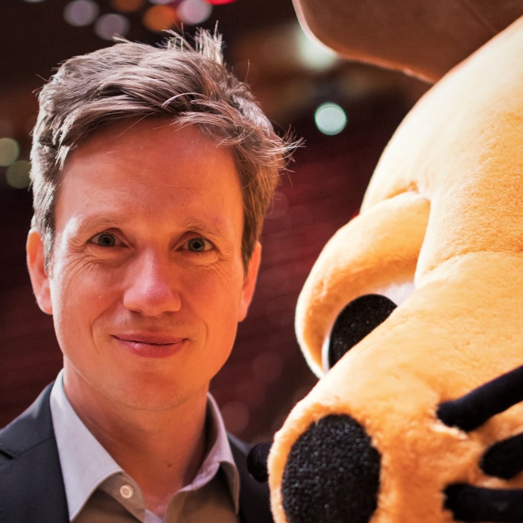 Man with short light brown hair and wearing a suit smiles next to a large yellow plush animal with black nose and whiskers.