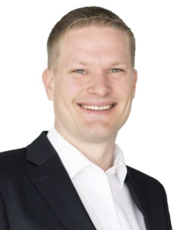 Portrait of a smiling man with short light brown hair wearing a black suit jacket and a white dress shirt against a white background.