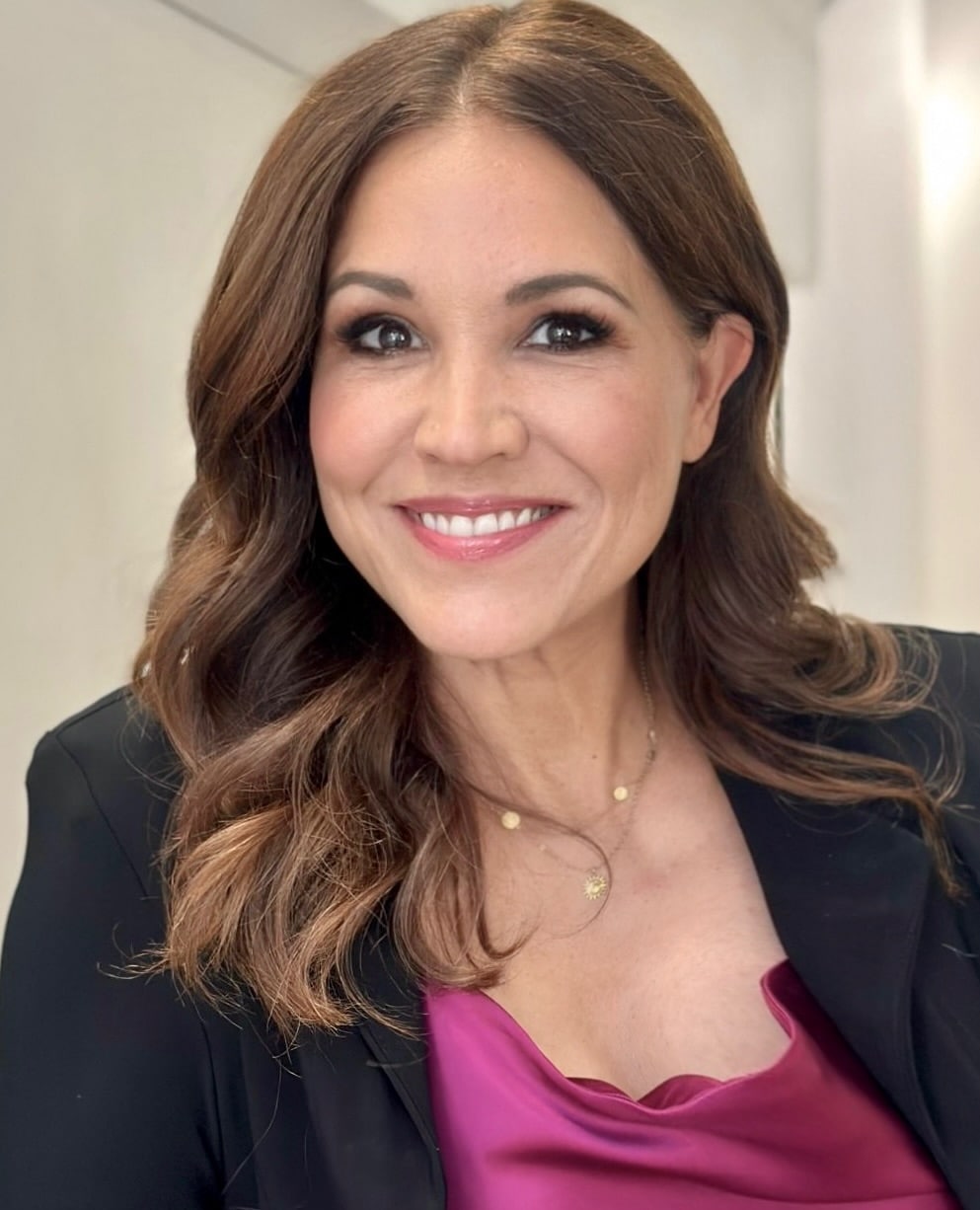 Smiling woman with light brown wavy hair wearing a black blazer and a magenta satin top, with a gold necklace featuring a sun pendant, against a neutral background.