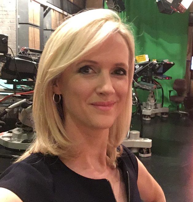 Blonde woman with shoulder-length hair wearing hoop earrings and a black top, smiling slightly in a studio with camera equipment and green screen in the background.