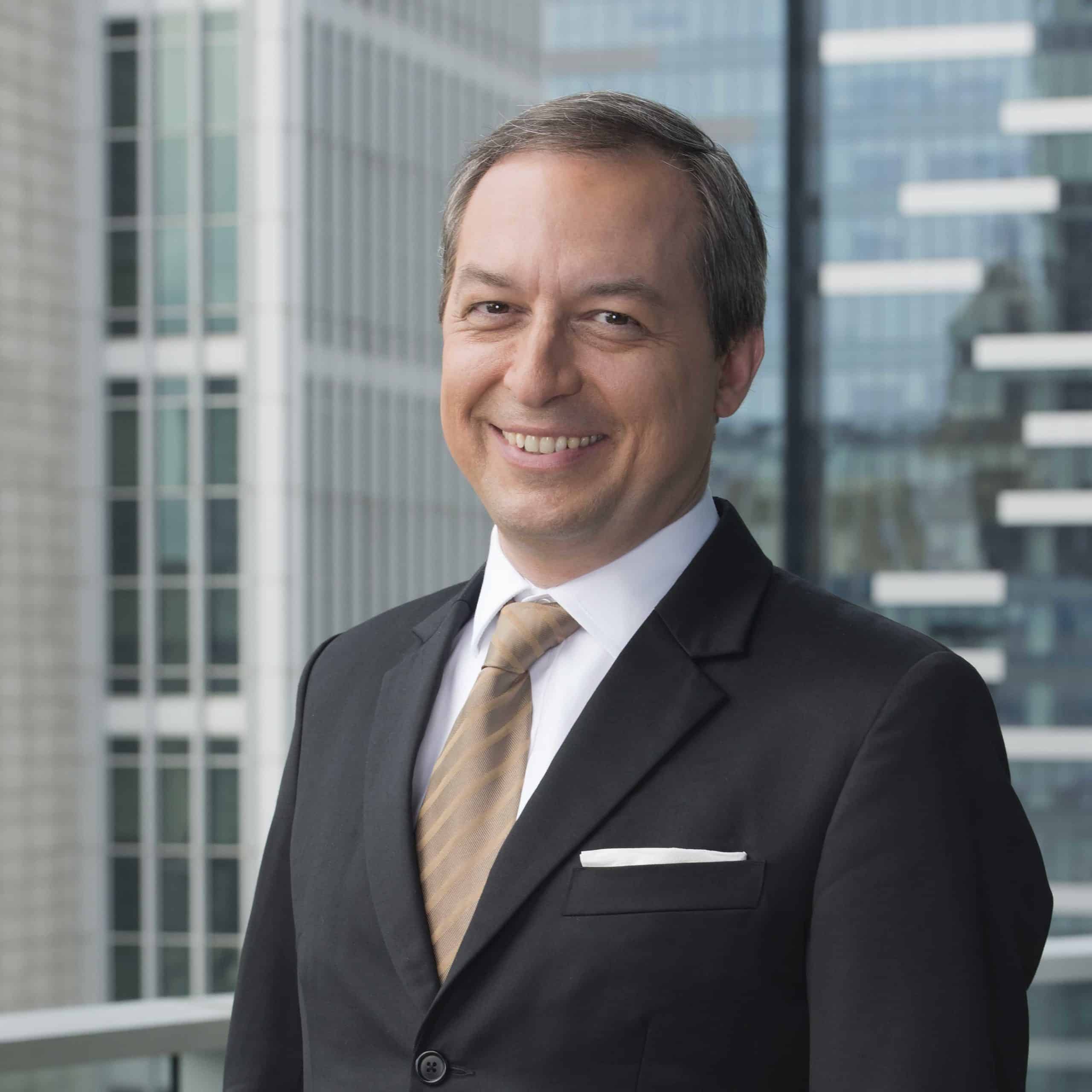 Smiling middle-aged man in a black suit, white dress shirt, and light brown striped tie standing in front of tall office buildings with glass windows.