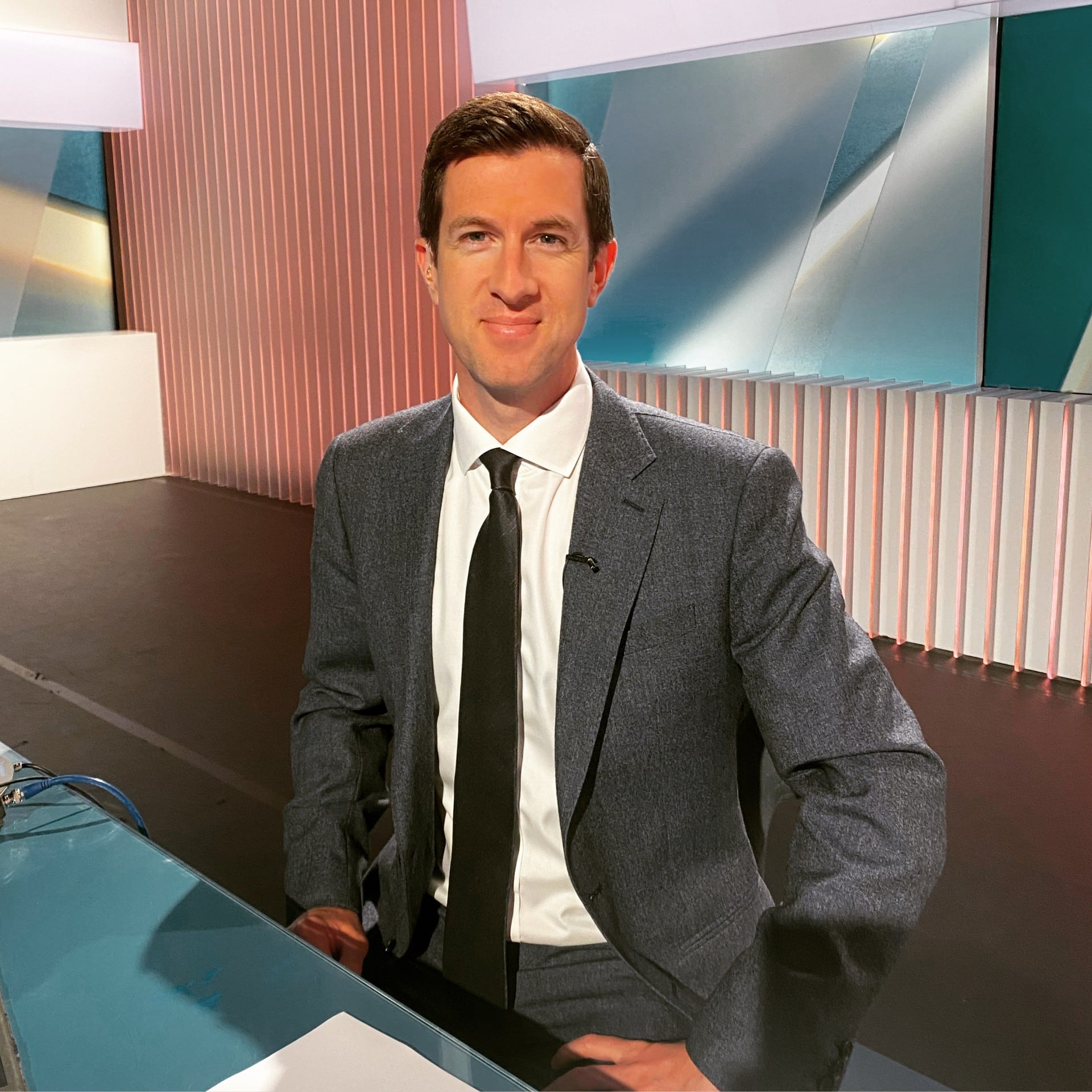 A man with short brown hair wearing a gray suit, white shirt, and black tie sits at a glass table in a modern studio with colorful background panels.