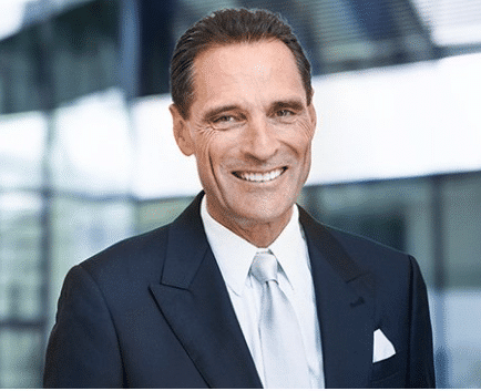 Portrait of a smiling middle-aged man with dark hair wearing a dark suit, white shirt, and white tie, standing in a modern office background.