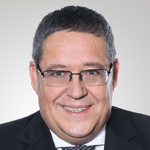 Portrait of a middle-aged man with short black hair, glasses, and a dark suit smiling against a plain light background.