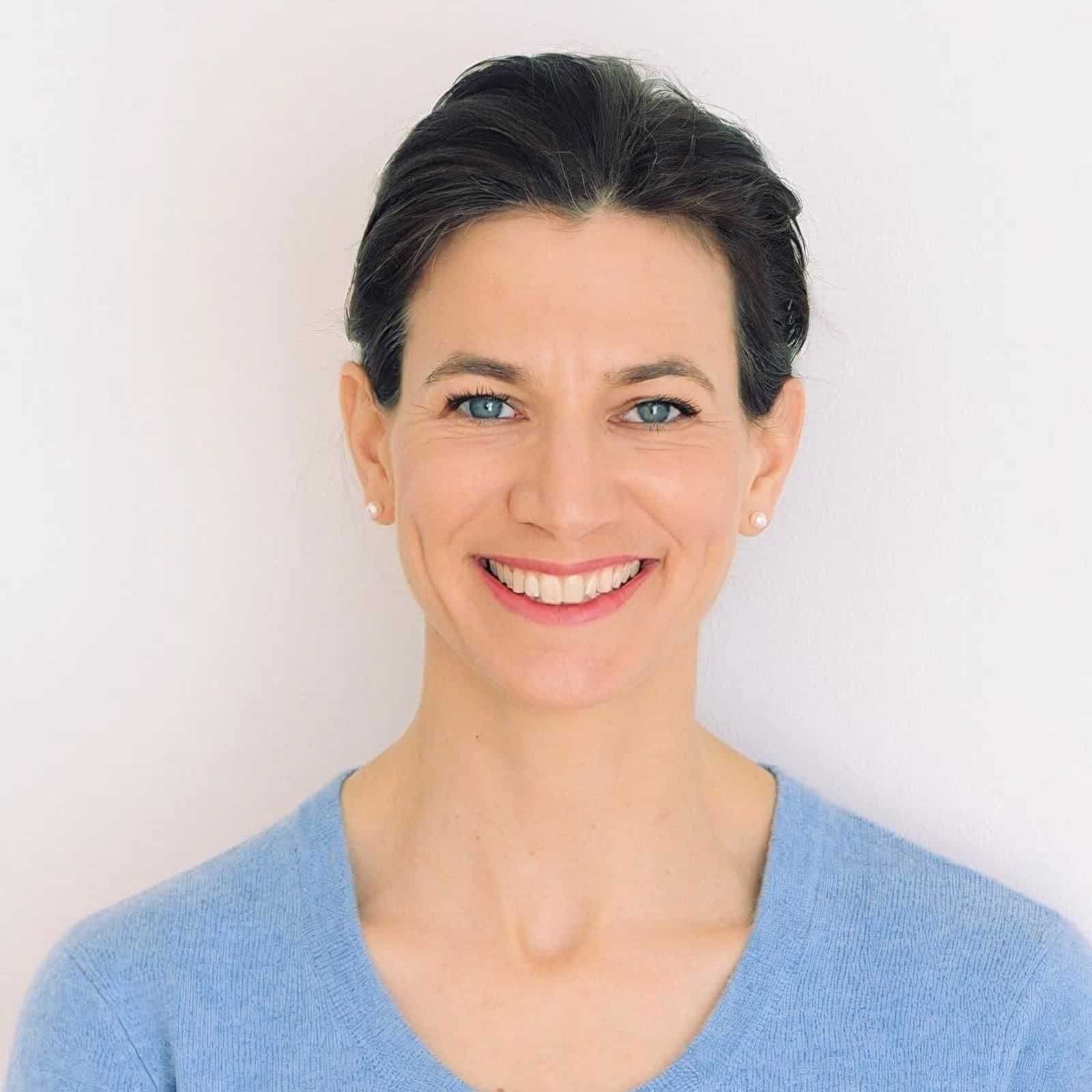 Smiling woman with dark hair pulled back, wearing small pearl earrings and a light blue sweater, against a plain white background.