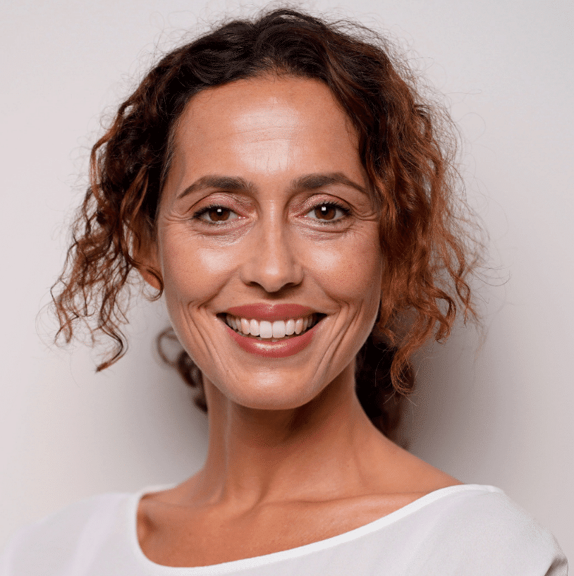 Smiling middle-aged woman with curly brown hair wearing a white top against a plain light background.