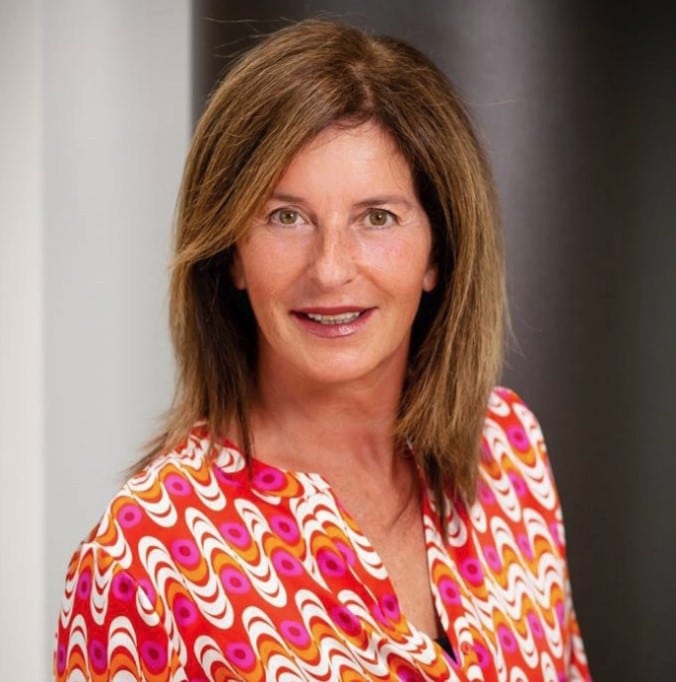 Smiling woman with shoulder-length light brown hair, wearing a colorful red, pink, orange, and white patterned top, standing against a neutral background.