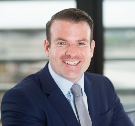 Smiling man in a dark blue suit, light blue shirt, and gray tie sitting indoors with blurred windows in the background.