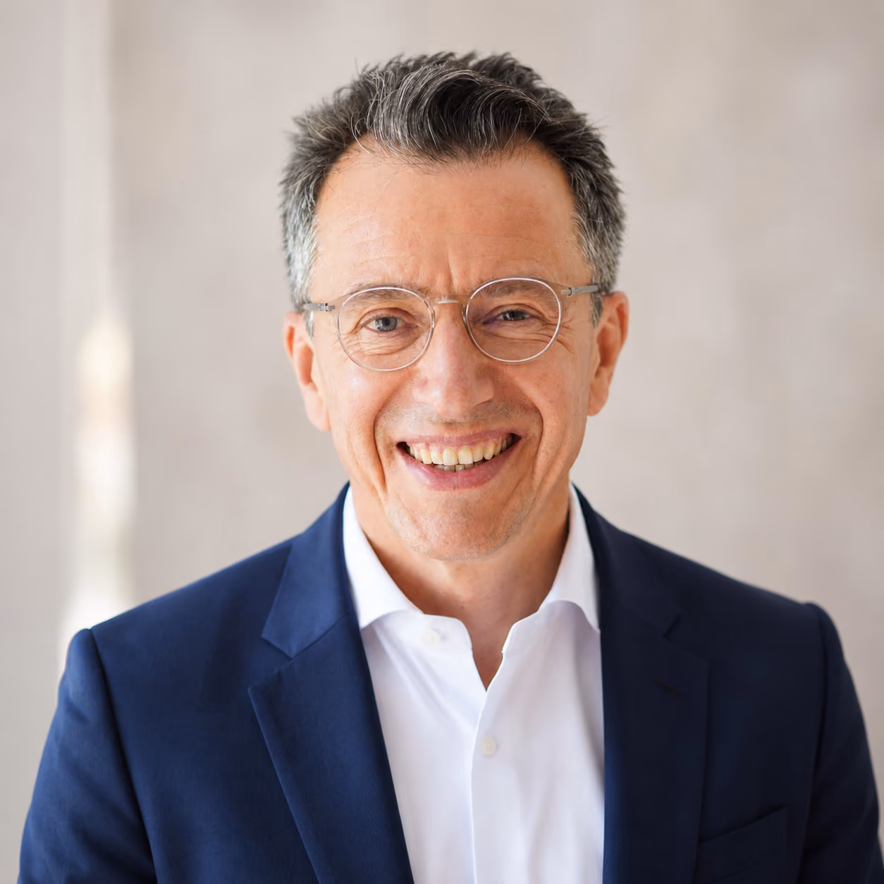 Smiling middle-aged man with short salt-and-pepper hair, wearing round glasses, a white shirt, and a dark blue blazer, standing against a neutral blurred background.