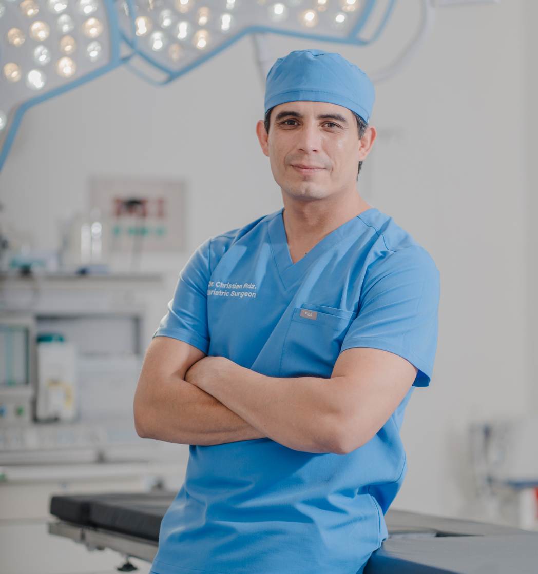 Dr, Christian in blue scrubs and cap standing with arms crossed in an operating room with surgical lights overhead.