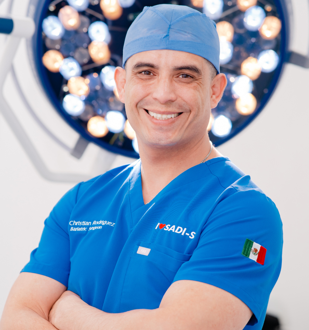 Dr, Christian bariatric surgeon in blue scrubs and cap, standing with arms crossed in a surgical room with bright round surgical lights behind him.
