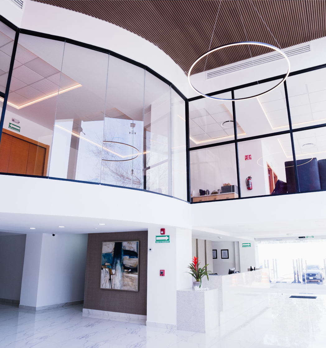 Modern lobby with white marble floors, glass-walled upper level, circular light fixtures, a reception desk with a red flower in a vase, and abstract artwork on a textured wall.
