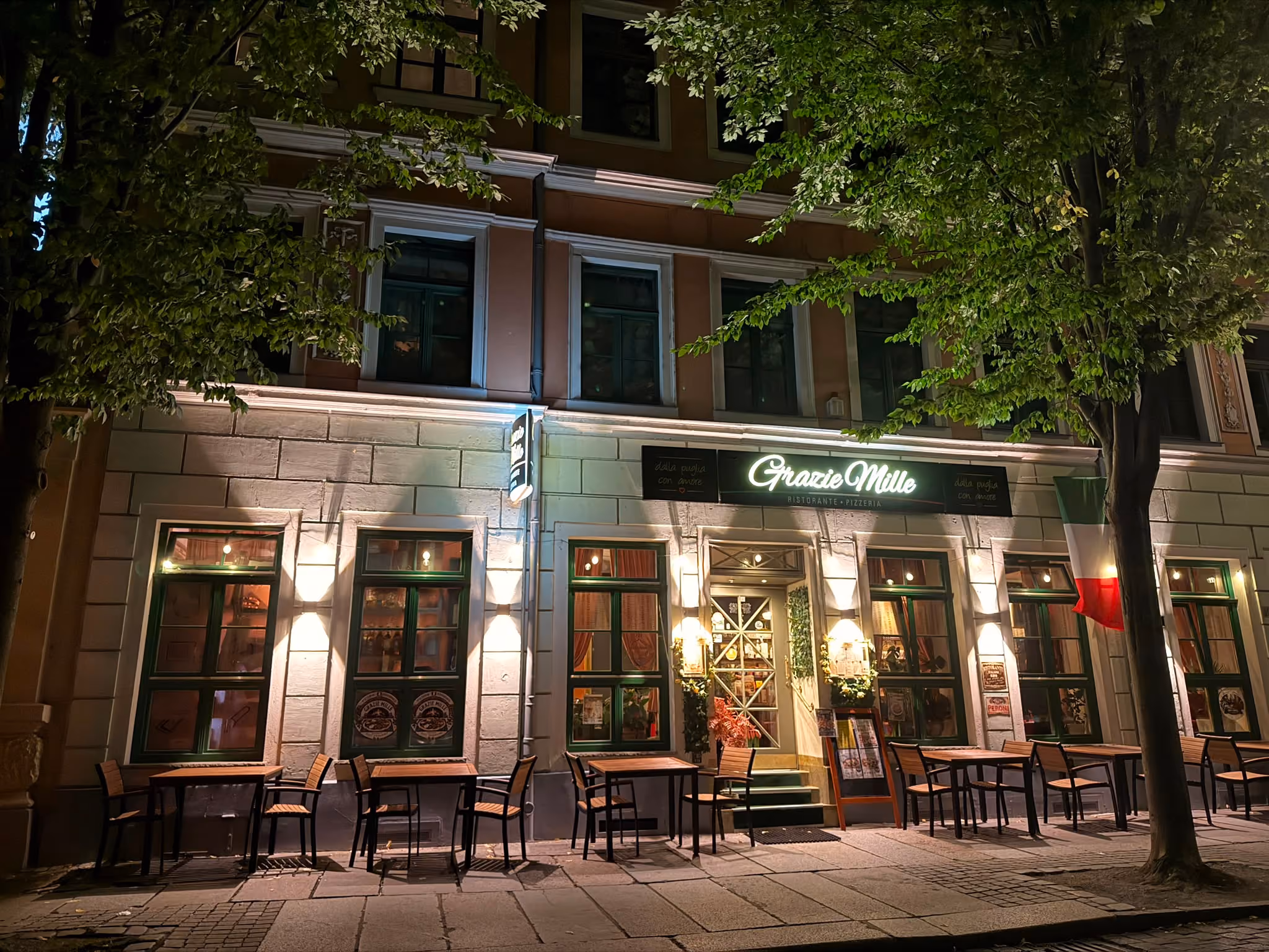 Beleuchtetes Restaurant mit dem Namen Grazie Mille an einem Baum gesäumten Bürgersteig bei Nacht mit mehreren Tischen und Stühlen im Außenbereich.