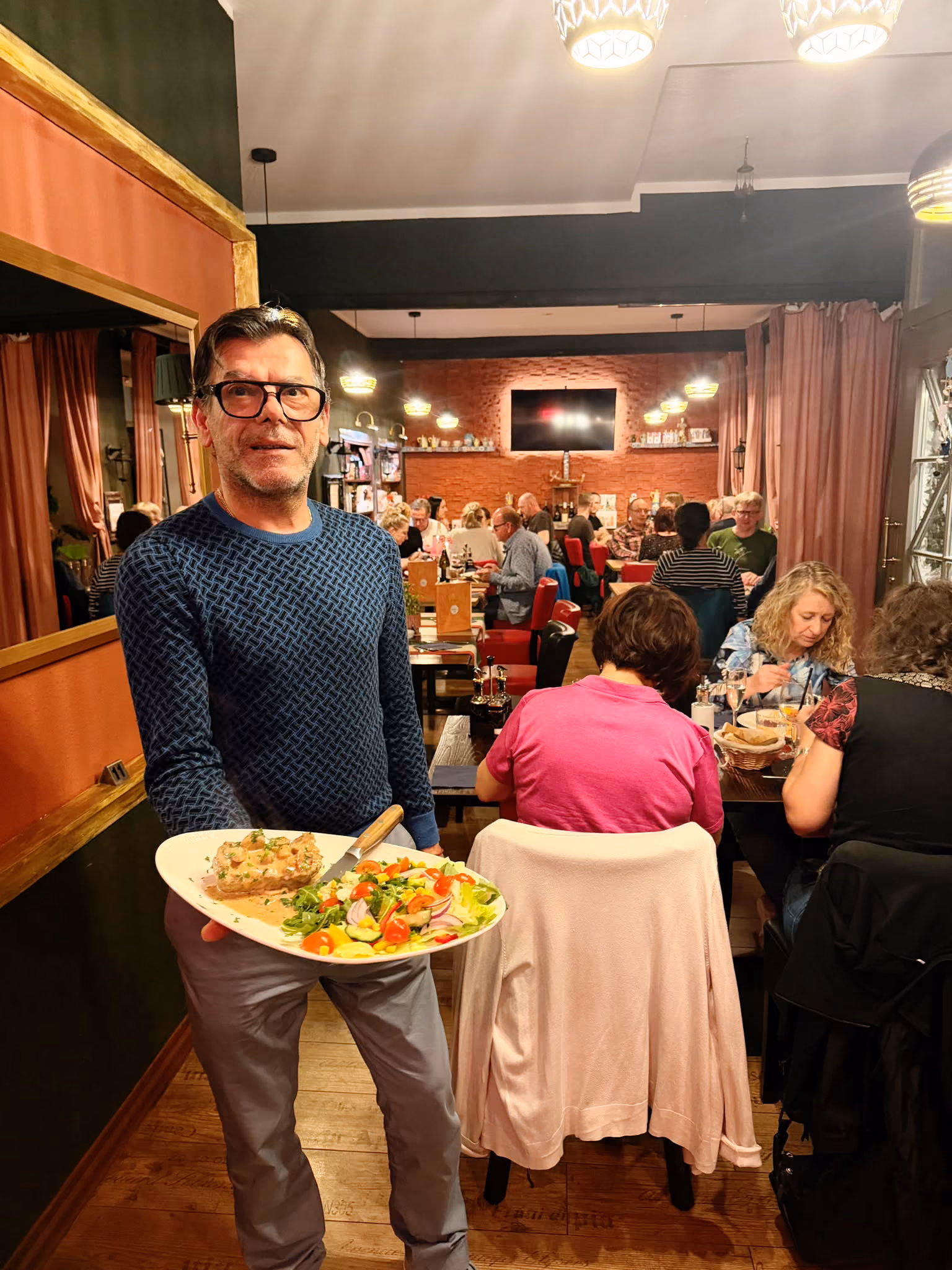 Ein Mann mit Brille und blauem Pullover hält einen Teller mit einem Salat und einer Scheibe Brot in einem belebten Restaurant mit mehreren Gästen im Hintergrund.