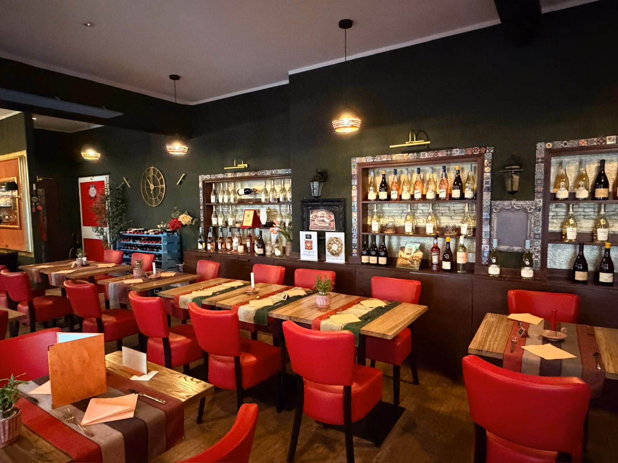 Innenraum eines Restaurants mit mehreren Holztischen und roten gepolsterten Stühlen, dekoriert mit Flaschen und Glasgefäßen in Regalen an der dunklen Wand, warme Beleuchtung.