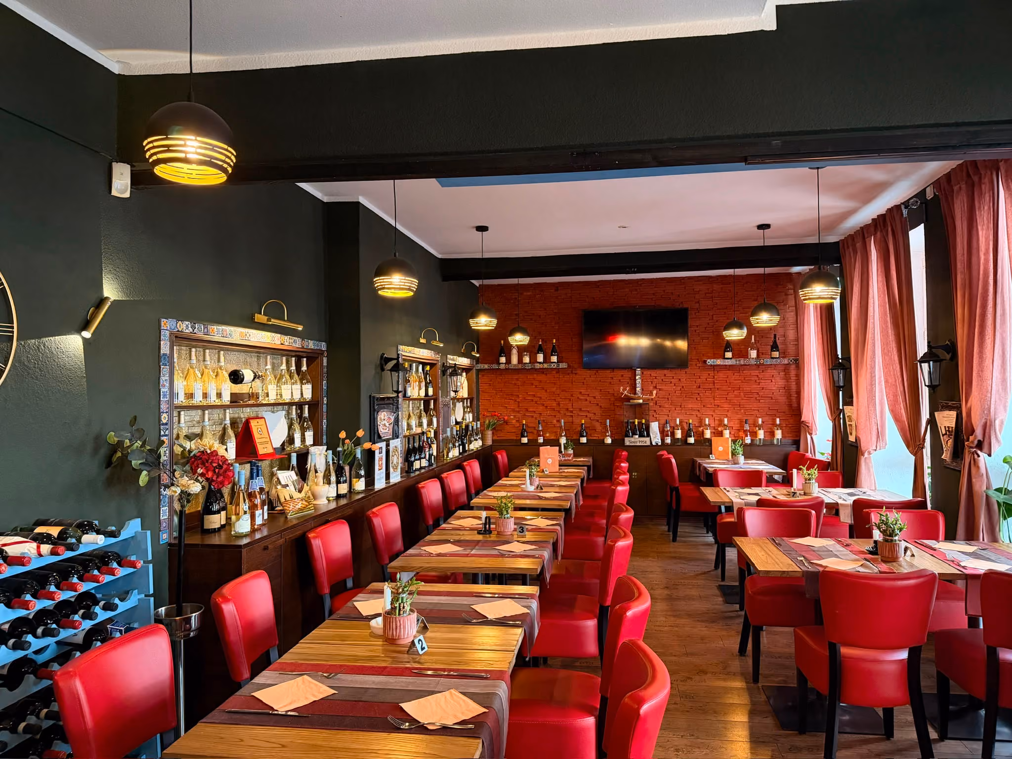 Modernes Restaurant mit roten Stühlen und Holztischen, an denen kleine Pflanzen als Dekoration stehen, mit Weinflaschenregal an der linken Wand und einem Fernseher an der rot gestrichenen Rückwand, sowie großen Fenstern mit roten Vorhängen auf der rechten Seite.