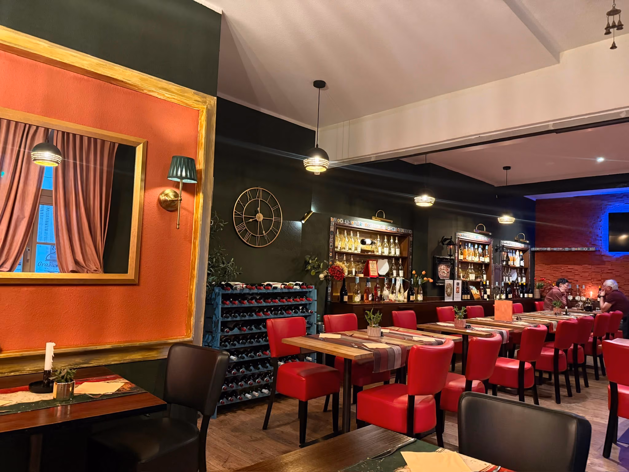 Innenraum eines Restaurants mit roten Stühlen und Holztischen, an der Wand große Weingestelle, eine Uhr und dekorative Regale mit Flaschen, gedämpfte Beleuchtung und zwei Personen, die am Tisch sitzen.