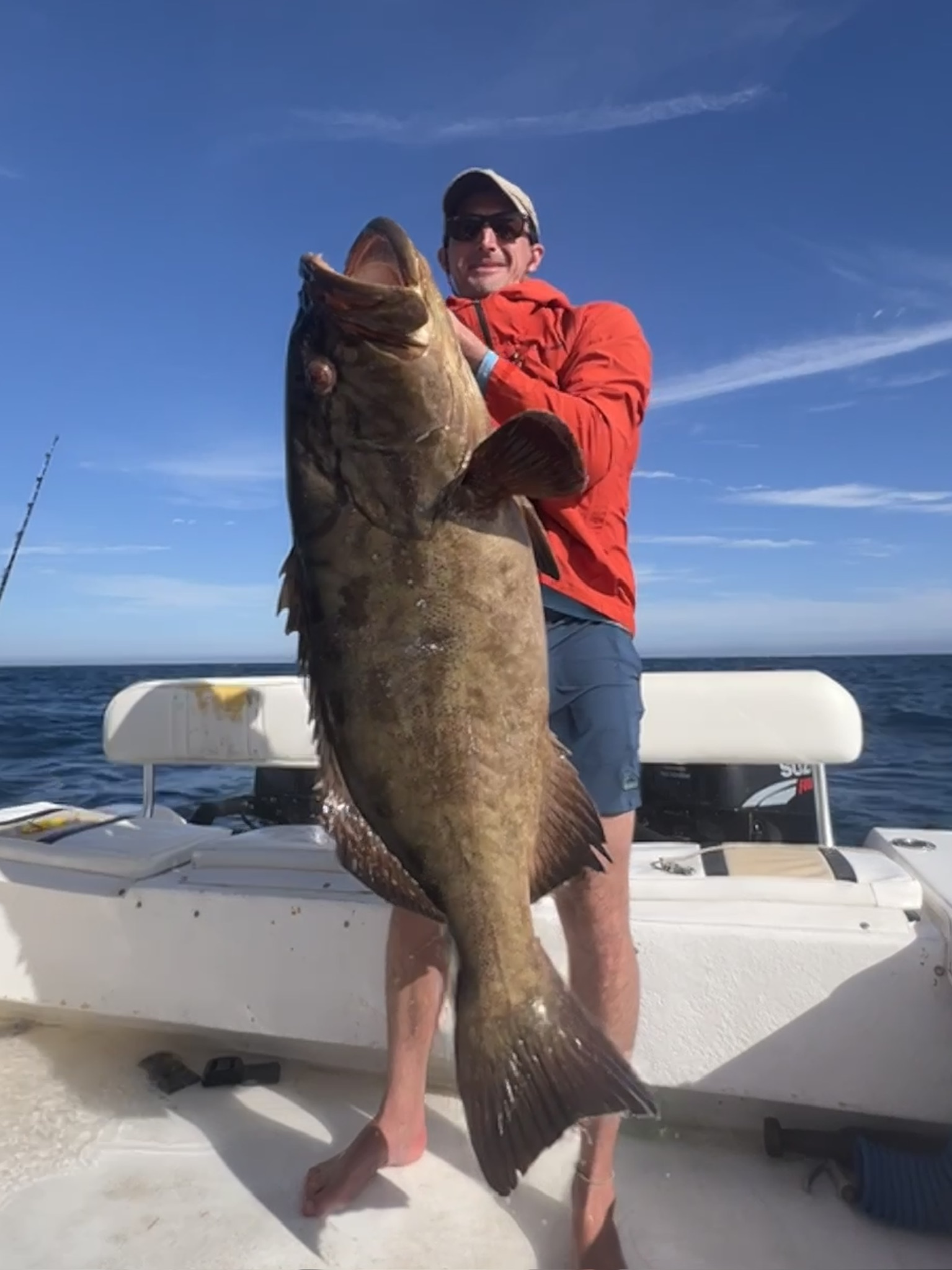 A man in a red jacket holds a very large brown grouper upright on the deck of a boat offshore.
