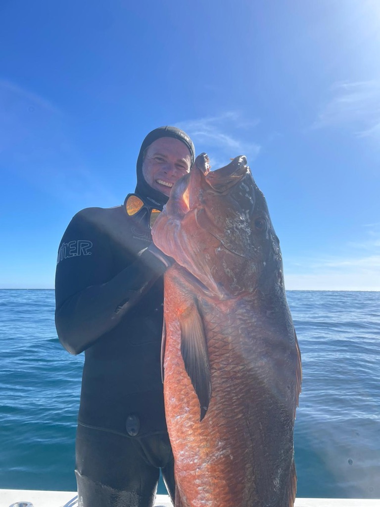 A freediver in a black wetsuit holds up a very large red snapper on the deck of a boat under a bright sky.