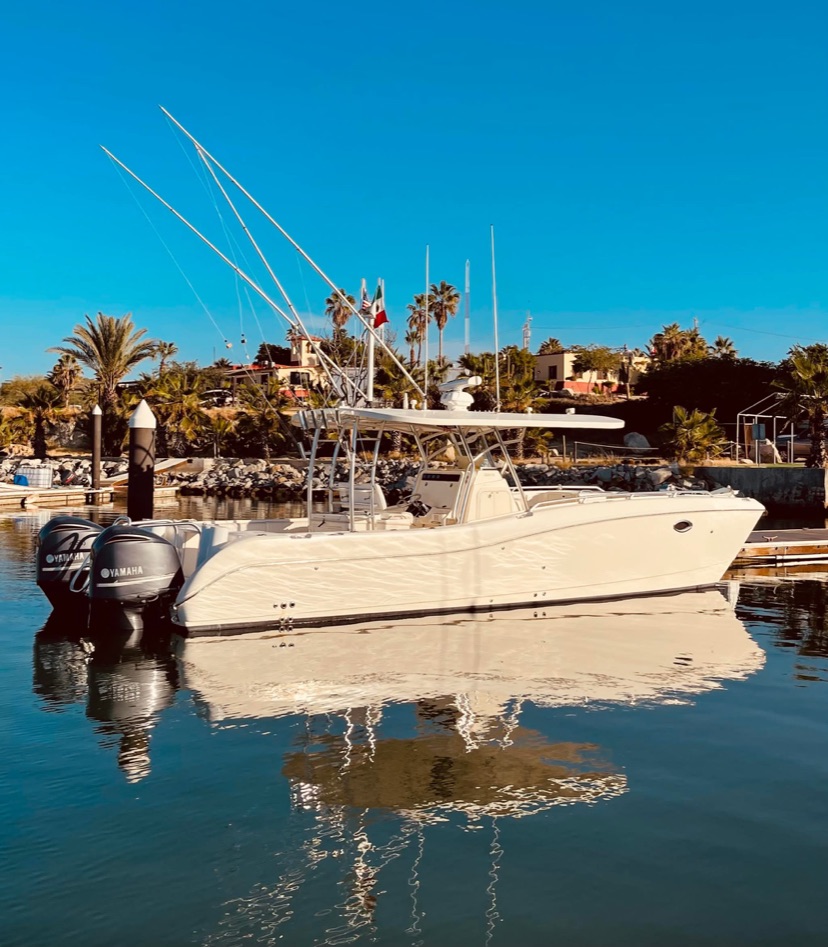 A white center console sportfishing boat with outriggers and triple outboards sits mirrored in still marina water, with palms and a Mexican flag behind.