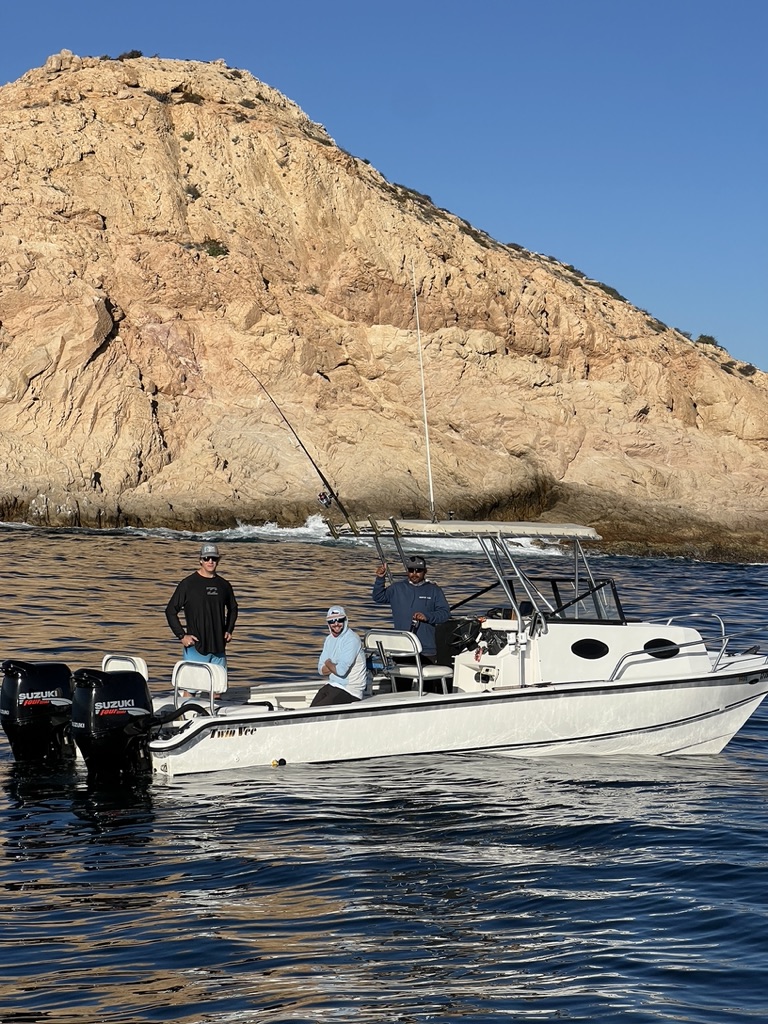 Three anglers stand aboard a white centre-console fishing boat on calm blue water below a large sunlit granite headland.
