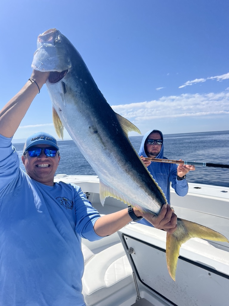 A smiling man in a blue sun shirt holds up a large silver fish with bright yellow fins while a second angler stands behind him with a rod.