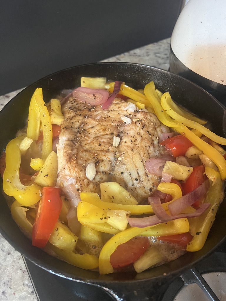 A seared white fish fillet simmering in a cast iron skillet with sliced yellow peppers, red onion and tomato.
