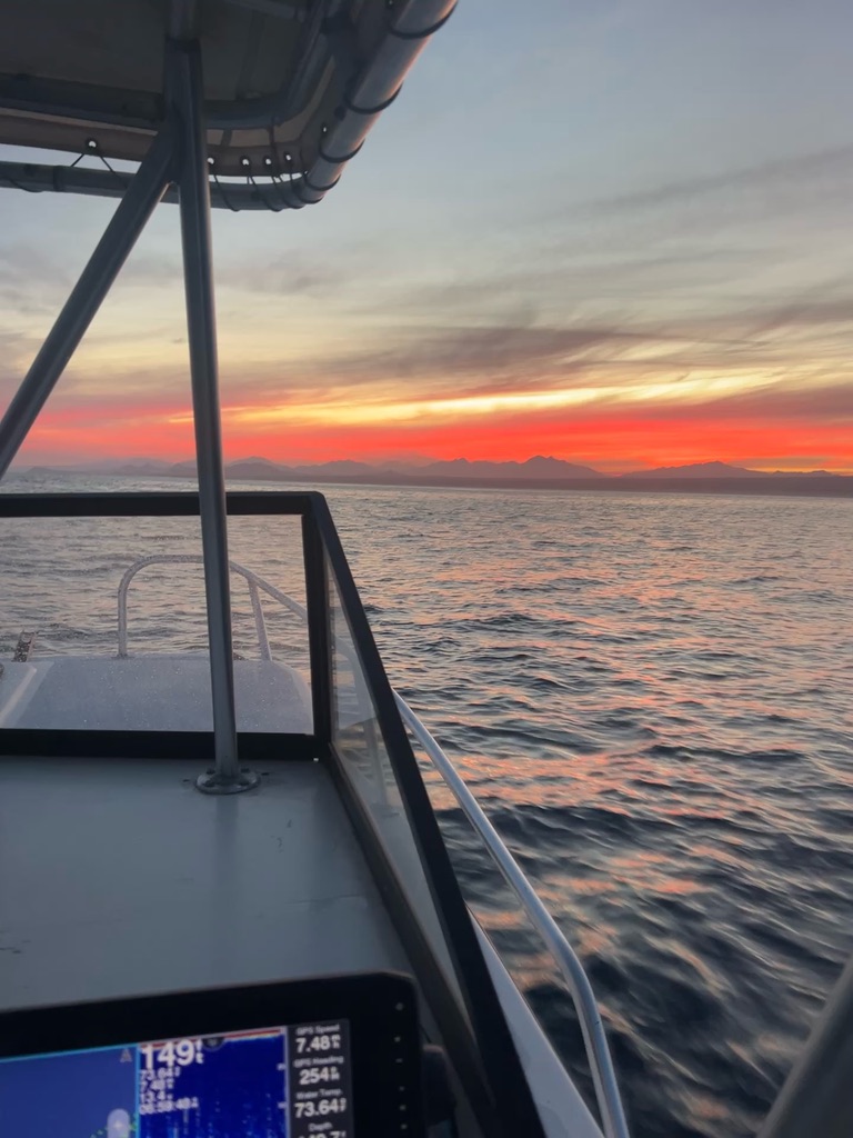 An orange sunrise burns over distant mountains seen past the rail and electronics of a moving boat.