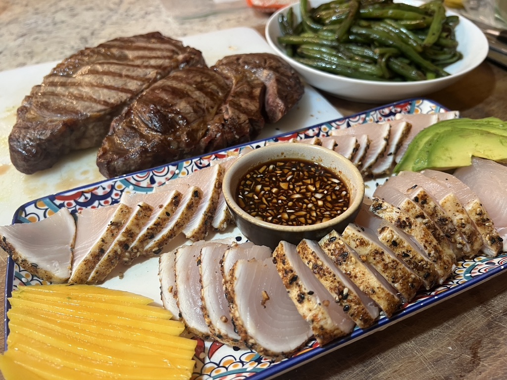 Grilled steaks on a board beside a platter of sesame-crusted seared tuna slices, sliced mango and a bowl of dipping sauce.