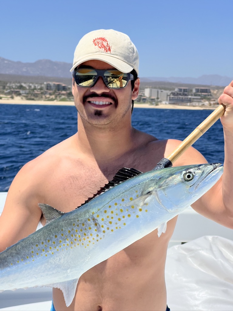 A shirtless man in a cap and sunglasses holds up a spotted silver mackerel on the boat with the coastline behind.