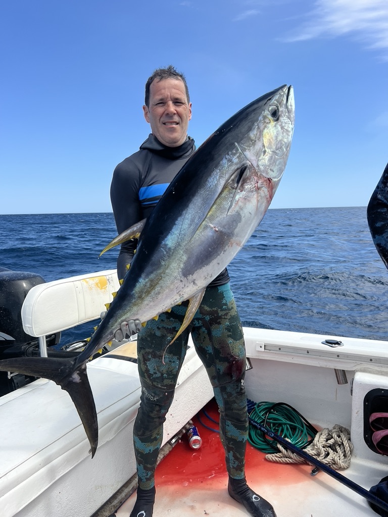A spearfisher in a camouflage wetsuit stands on the deck holding a large yellowfin tuna upright against his chest.