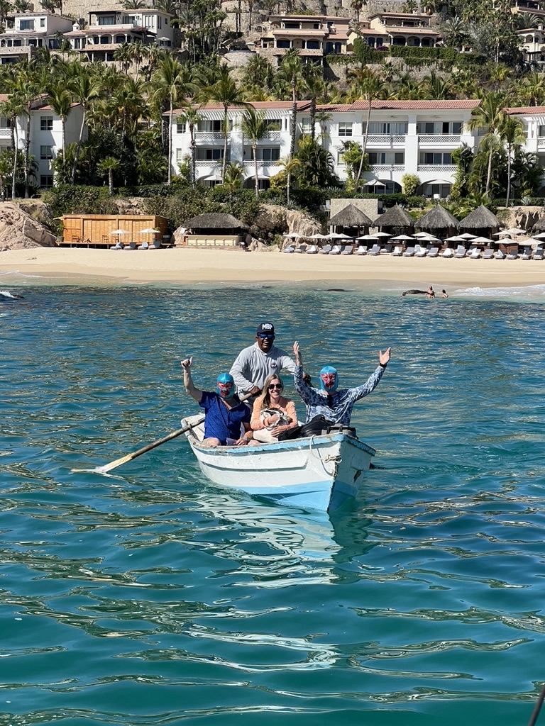 Four people wave from a small white skiff on turquoise water in front of a palm-lined beach resort and rows of thatched umbrellas.