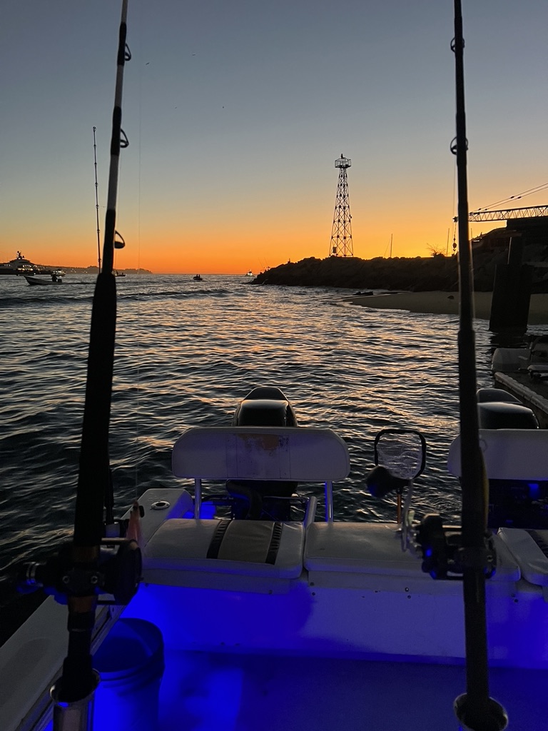Fishing rods stand in their holders on a boat lit by blue deck lights as the sun sets behind the harbour breakwater and its light tower.