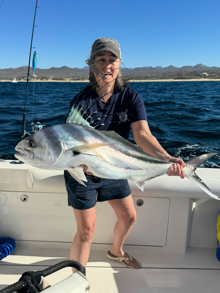 A woman holds a large roosterfish across her arms on the deck of a boat with the Baja coastline behind her.
