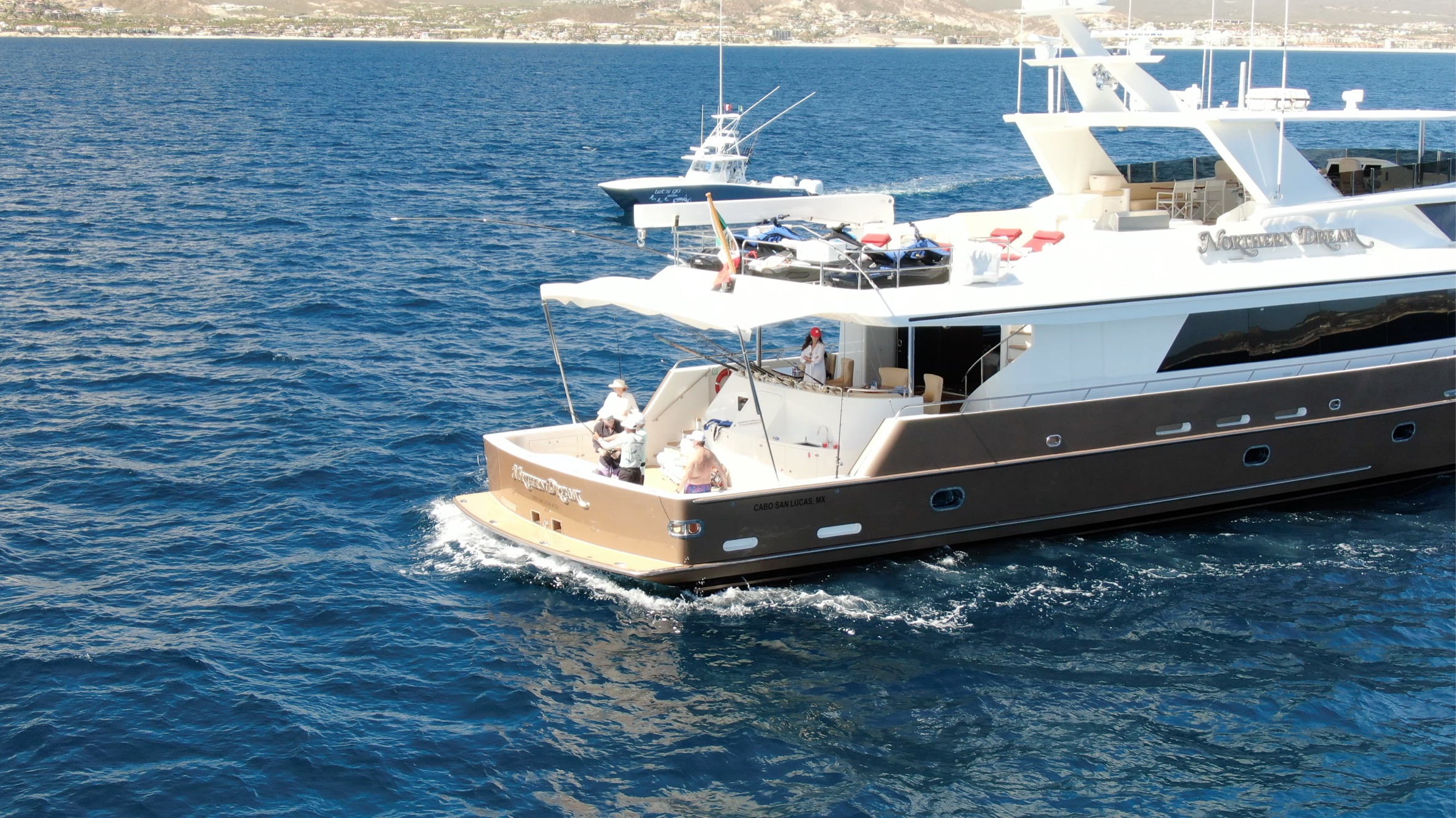 A large motor yacht with anglers fishing from its open stern cockpit, another sportfishing boat beyond it on deep blue water.