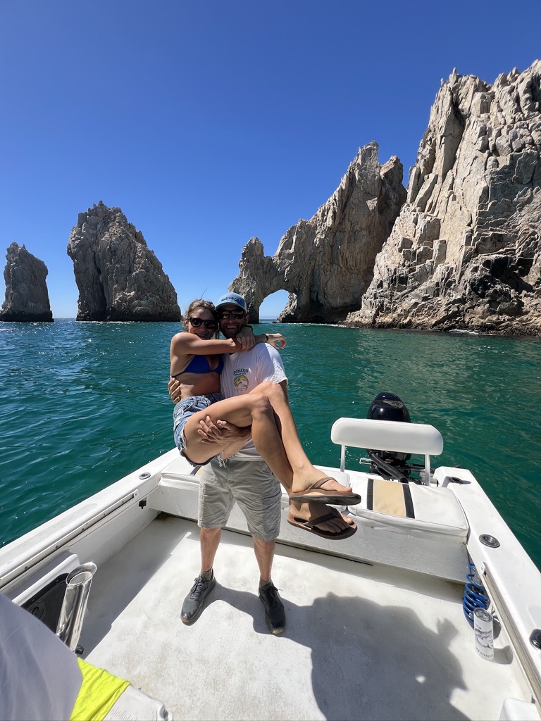 A man carries a woman in his arms on the bow of a small boat in front of the rock arch at Land's End in Cabo San Lucas.