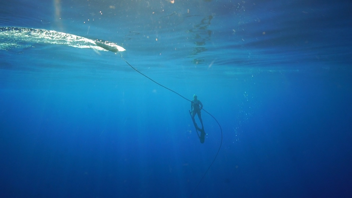 A freediver in long fins descends a weighted line into deep blue water with sunlight streaming down from the surface above.