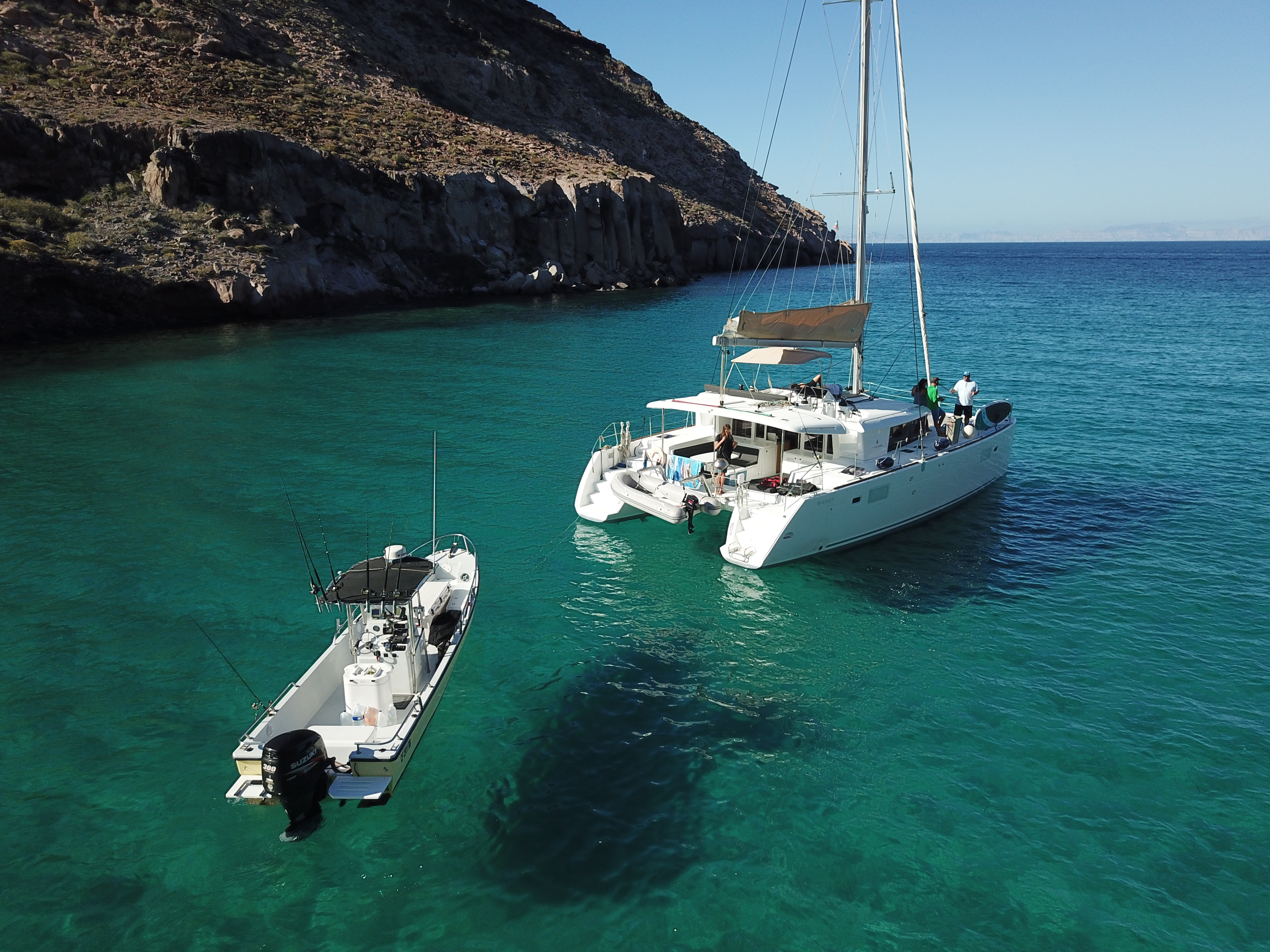 An aerial view of a sailing catamaran and a fishing panga anchored in clear green-blue water beneath an arid rocky ridge.