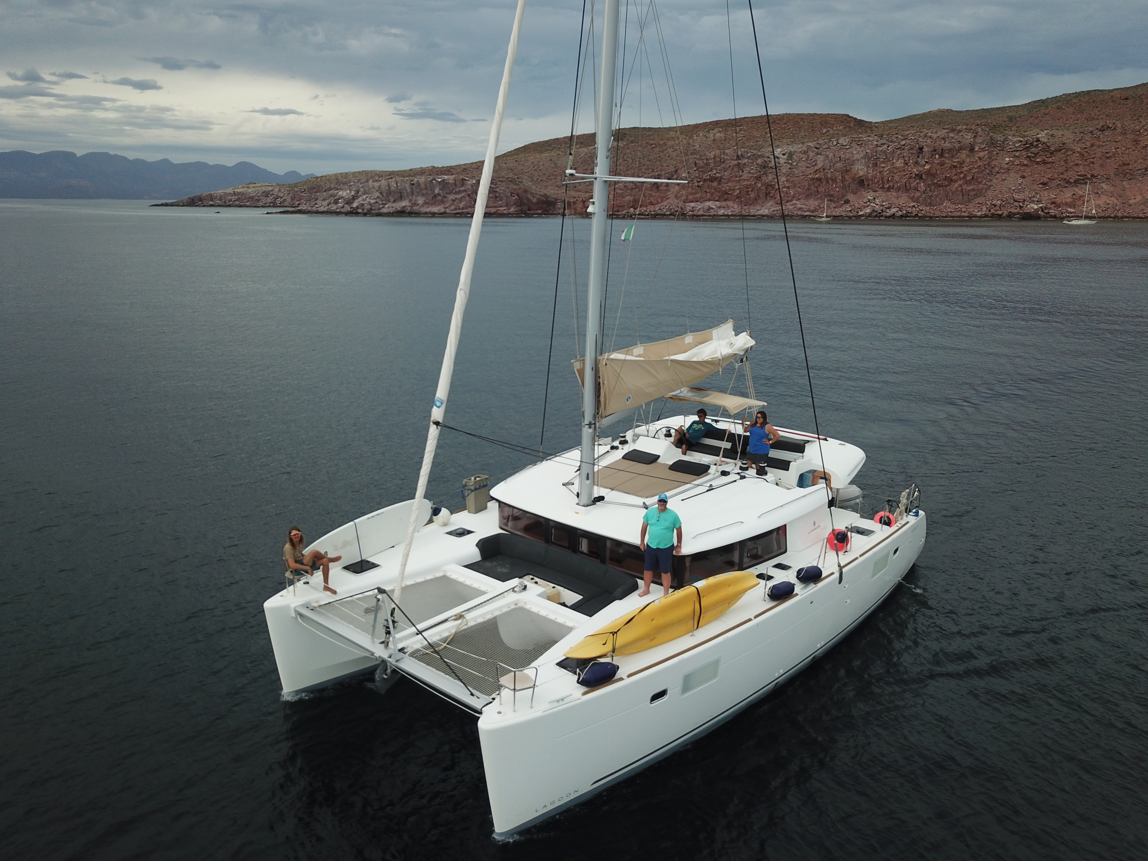 A sailing catamaran lies at anchor on calm water with several people relaxing on its deck and a yellow kayak stowed aft.