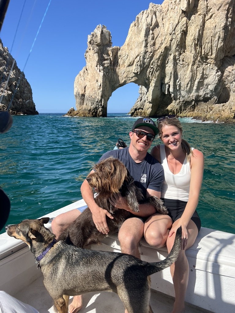 A couple sit with two dogs on the bow of a boat in front of the natural rock arch at Land's End.