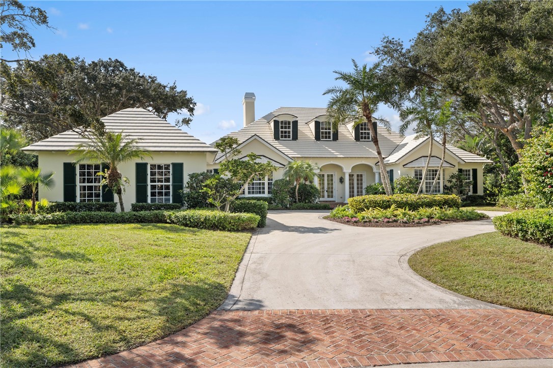 Stucco exterior residence with a tiled roof, featuring a circular driveway, mature landscaping, dark louvered shutters, and a prominent chimney