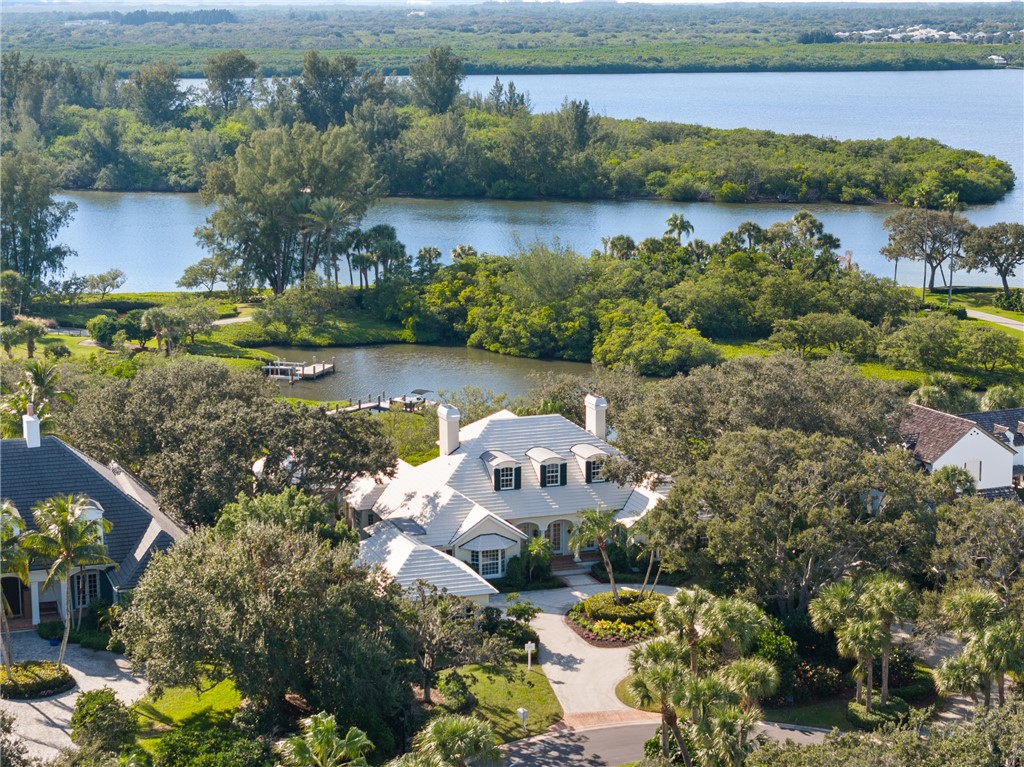 Expansive property featuring a waterfront setting with a private boat dock, lush tropical landscaping, mature trees, a circular driveway, and a multi-level home with a white tile roof and multiple dormer windows