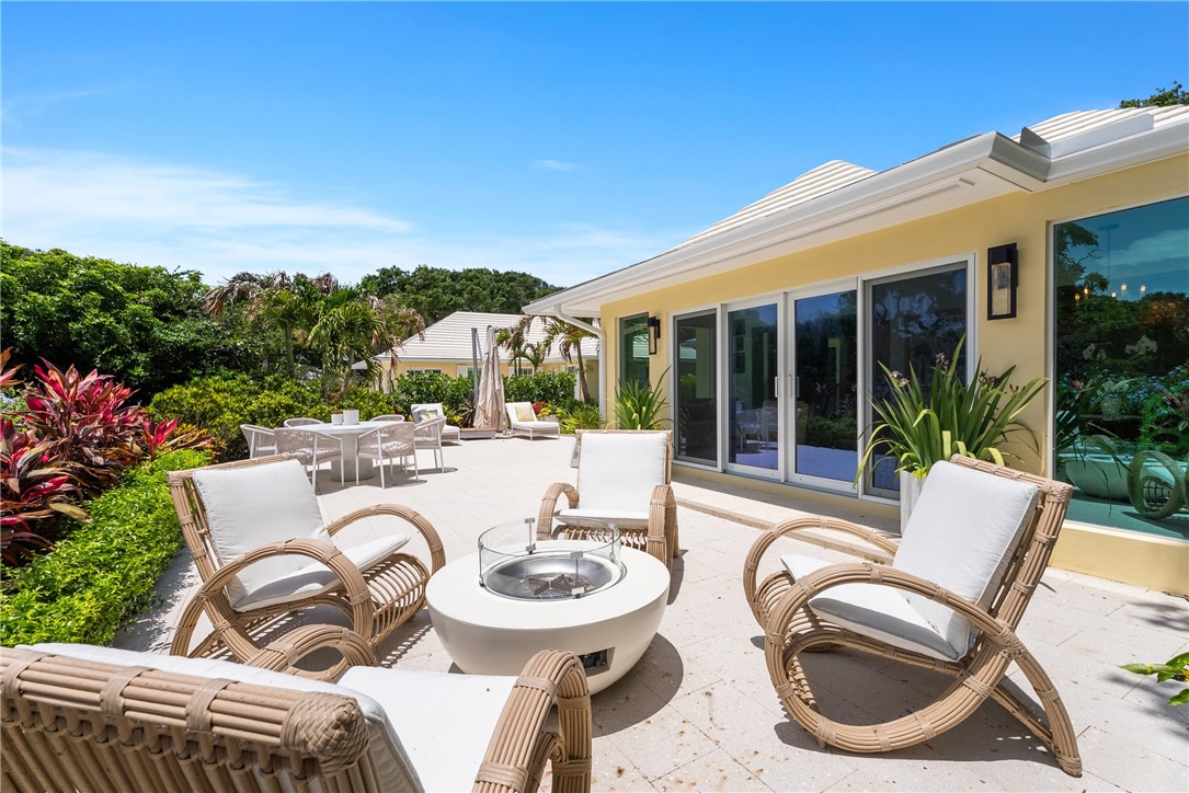 Expansive outdoor patio featuring a fire pit, mature tropical landscaping, multiple seating areas, and a light yellow exterior with large sliding glass doors