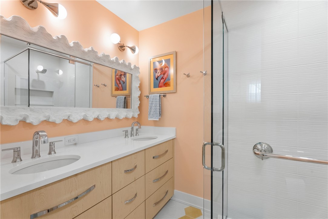 Dual vanity with undermount sinks and chrome fixtures, light wood cabinetry with curved hardware, and a glass-enclosed shower with white textured tile