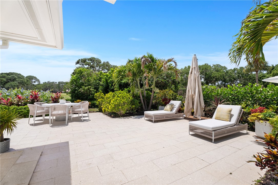 Expansive outdoor patio featuring light-colored paver flooring, lush tropical landscaping, and a retractable white awning