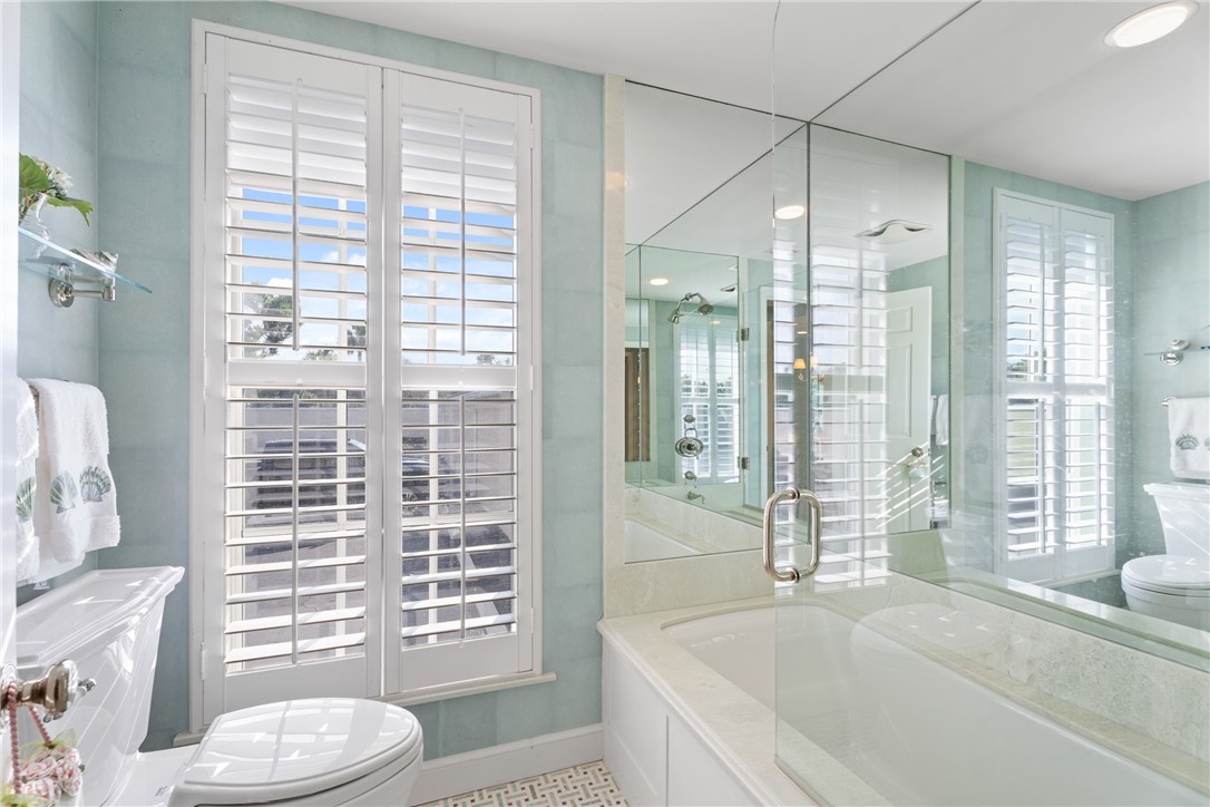 Bathroom featuring a built-in tub with a clear glass enclosure, full-height mirrored wall, and plantation shutters on dual windows