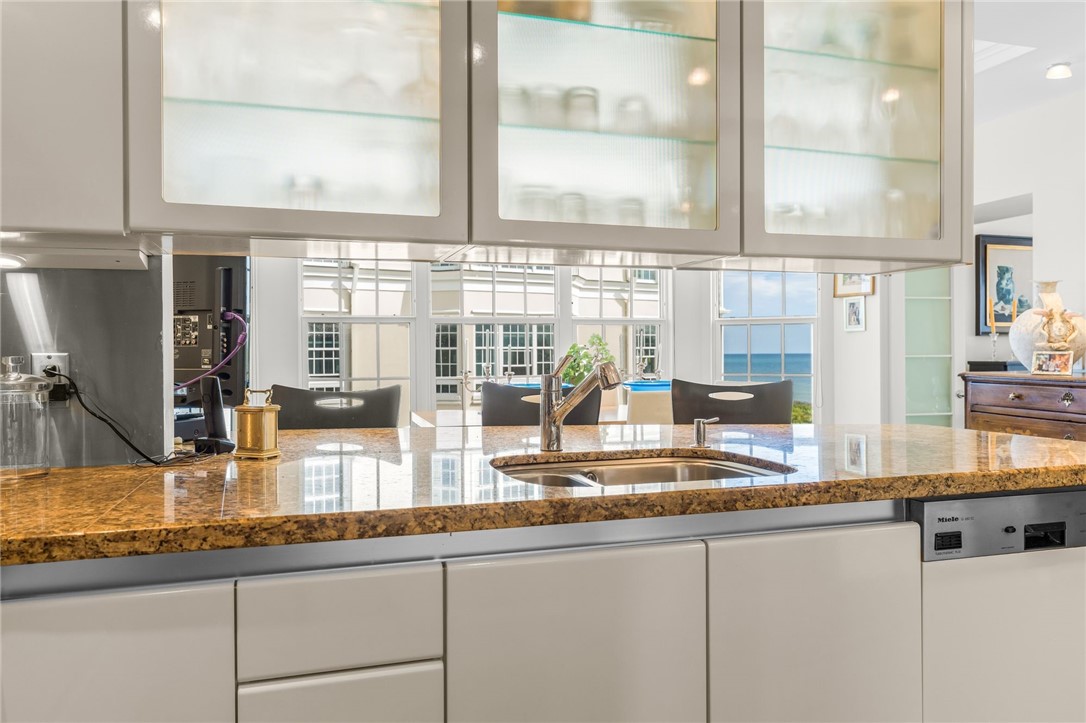 Contemporary kitchen featuring custom cabinetry with glass-front upper cabinets, warm-toned stone countertops, stainless steel appliances, and abundant natural light from expansive windows overlooking the grounds