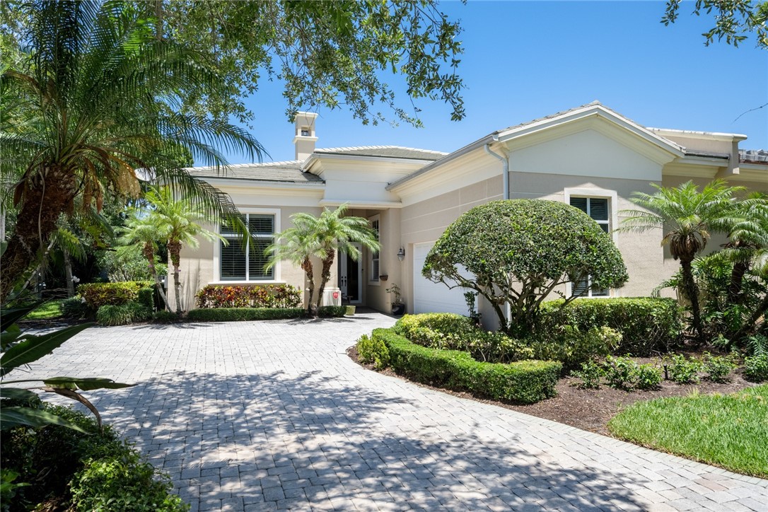 Paver driveway leading to a residence with a stucco exterior, multiple windows, and a prominent chimney feature