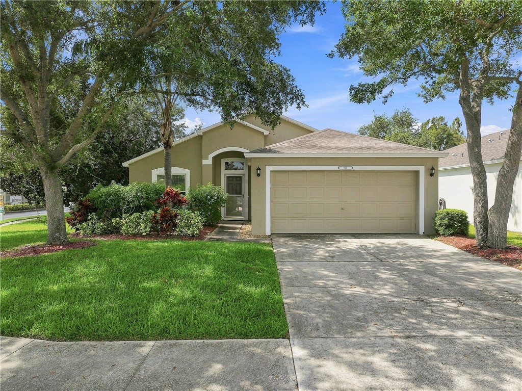 Single-story residence featuring a tan exterior with arched entryway, two-car garage with paneled door, and mature trees flanking the property