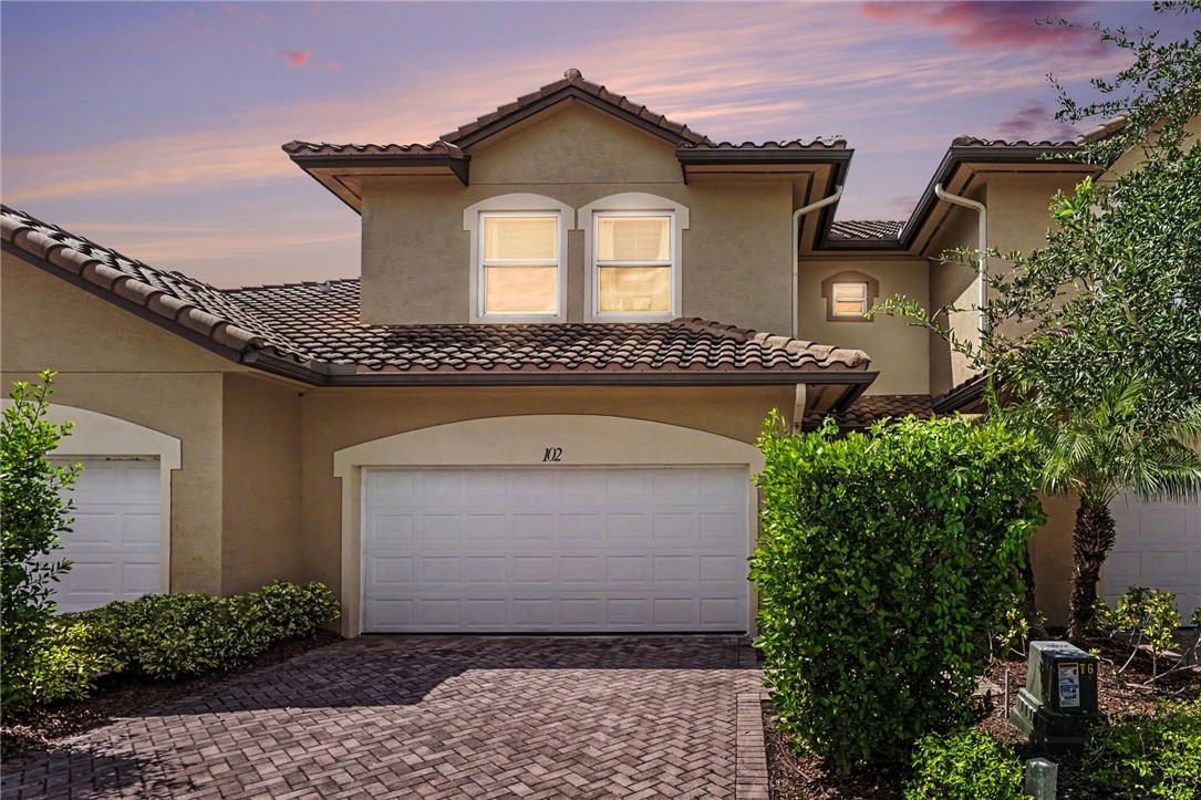 Stucco exterior with a barrel tile roof, arched garage entries, a paver driveway, and mature landscaping